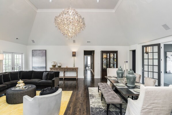 A spacious living room with a dark wood floor, a large chandelier, a black sofa, a gray armchair, and a dining area with a table set for four.