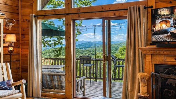Interieur van een houten chalet met een open haard, een eethoek en een schuifdeur die uitkomt op een terras met uitzicht op de bergen.