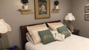A neatly made bed with a digital clock, a framed painting, and decorative plants on wall-mounted shelves.