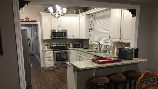 A kitchen with white cabinets, a microwave, an oven, and a coffee maker. There are three stools and a bar counter.