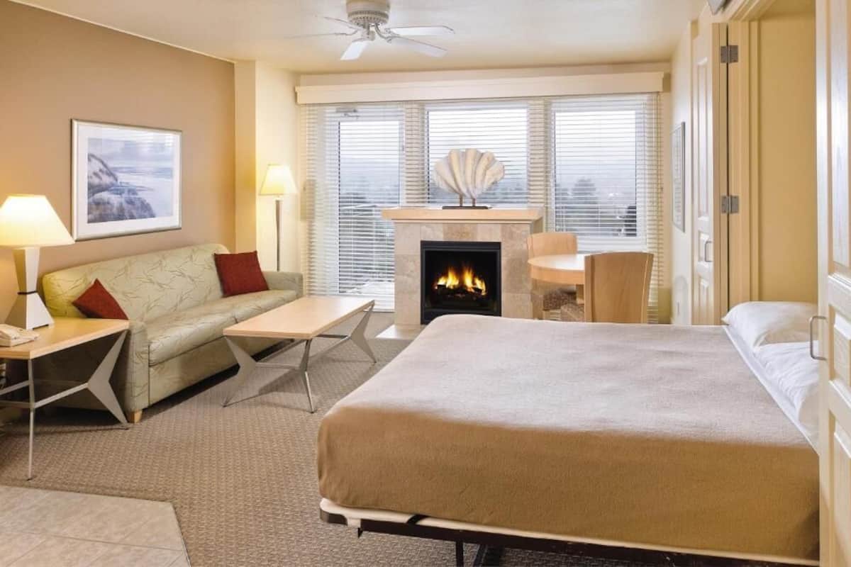 Habitación de hotel con cama, sofá, mesa, chimenea y vista a la ciudad a través de grandes ventanales.