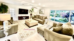A modern living room with a sofa, armchairs, a coffee table, and a TV. There is a window with a view of greenery outside.