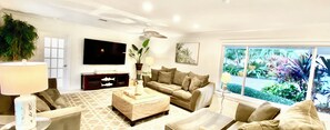Ein modernes Wohnzimmer mit Sofa, Sesseln, Couchtisch und Fernseher. Ein Fenster bietet Blick auf grüne Natur.