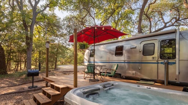 Ein Whirlpool mit einer Holterrasse, ein überdachter Picknicktisch und ein Wohnwagen in einer bewaldeten Gegend.