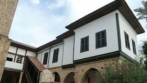 Un bâtiment historique aux murs blancs et aux encadrements de fenêtres noirs, doté d’une cour intérieure avec des arches en pierre et d’un escalier menant à un étage supérieur.