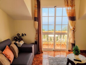 A living room with a sofa, a small table with a plant, and a view of a balcony with outdoor furniture. - Achada Hillside & Ocean View Apartment with Free WIFI in Arco da Calheta (Arco da Calheta)