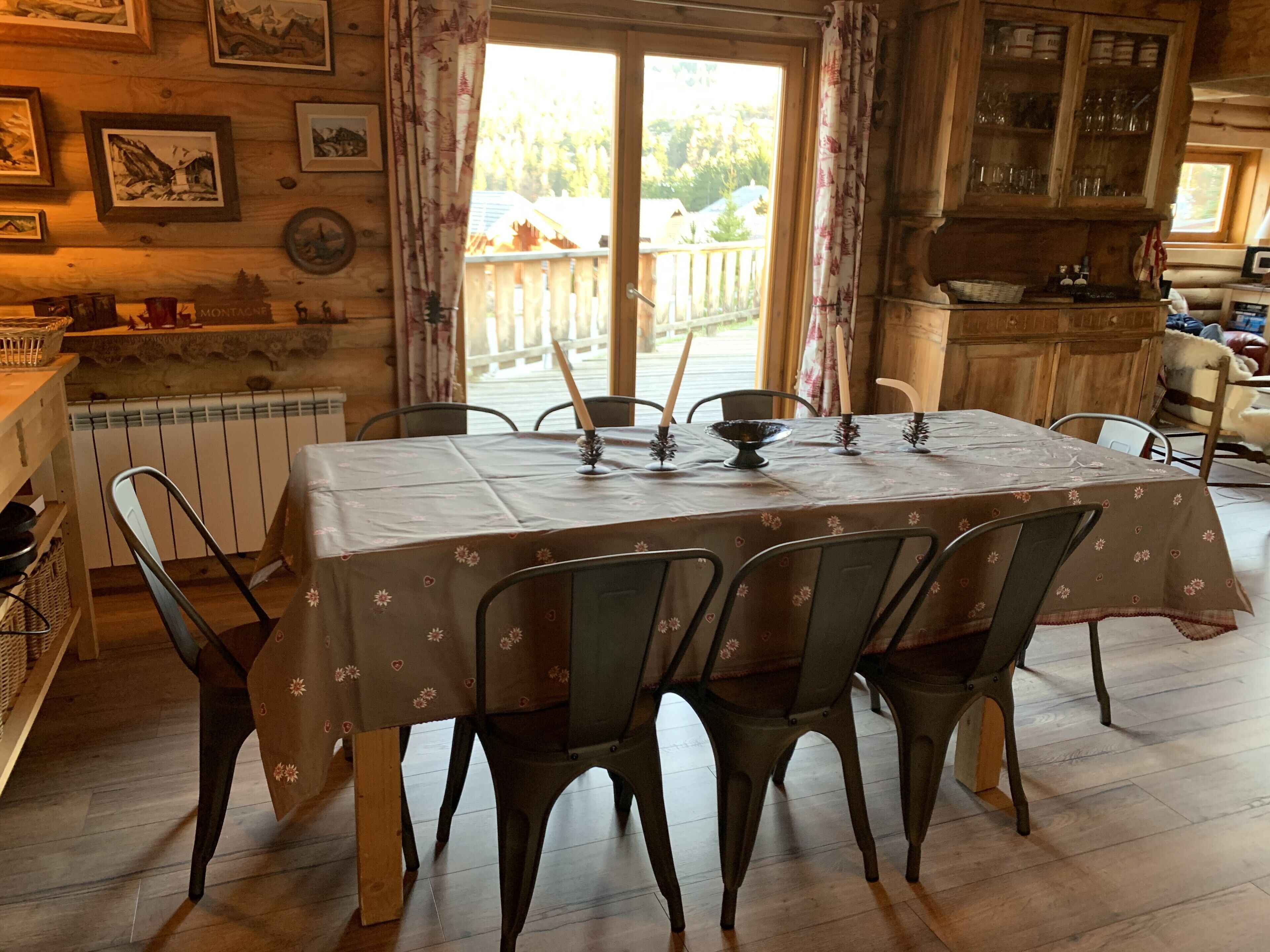 Ein Essbereich mit einem Tisch für vier Personen, umgeben von Stühlen. Ein Fenster mit Vorhängen ist sichtbar, und im Hintergrund befindet sich ein Holzschrank mit Gläsern.