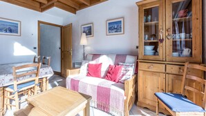 Een knus vertrek met een houten plafond, een bank met een geruit plaid, een eettafel met stoelen en houten kasten met glazen deuren.