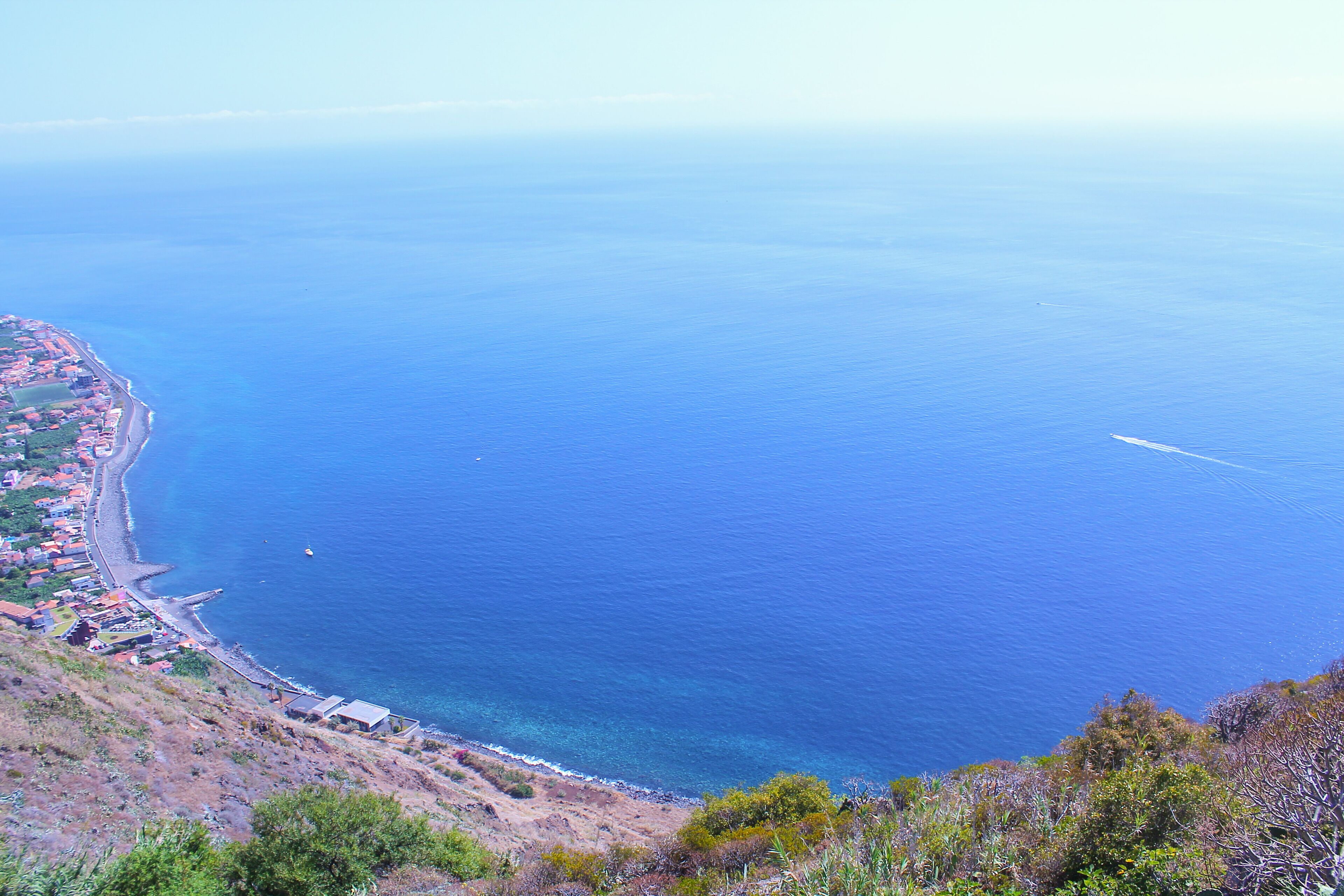 Vista costera con mar azul cristalino, un camino sinuoso a lo largo de la orilla y un paisaje montañoso con vegetación escasa.
