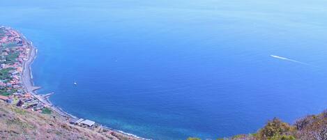Vista costera con mar azul cristalino, un camino sinuoso a lo largo de la orilla y un paisaje montañoso con vegetación escasa.