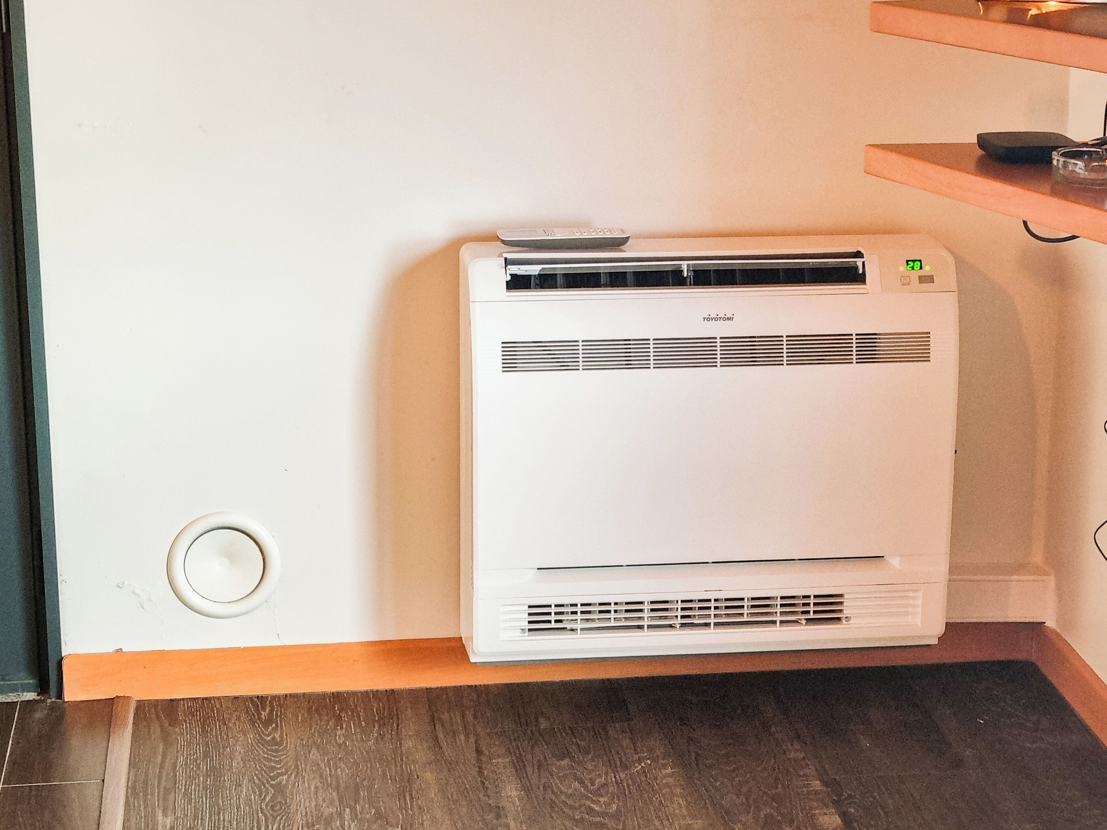 Een witte airconditioner in een hoek van een kamer met houten vloeren en een witte muur.