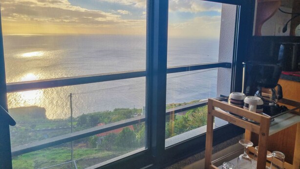 A room with large windows overlooking a body of water and greenery. There is a wooden chair and a coffee machine on a table. - Studio One | Ocean View | Perfect Budget Couple's Retreat with Free WIFI (Fajã da Ovelha)