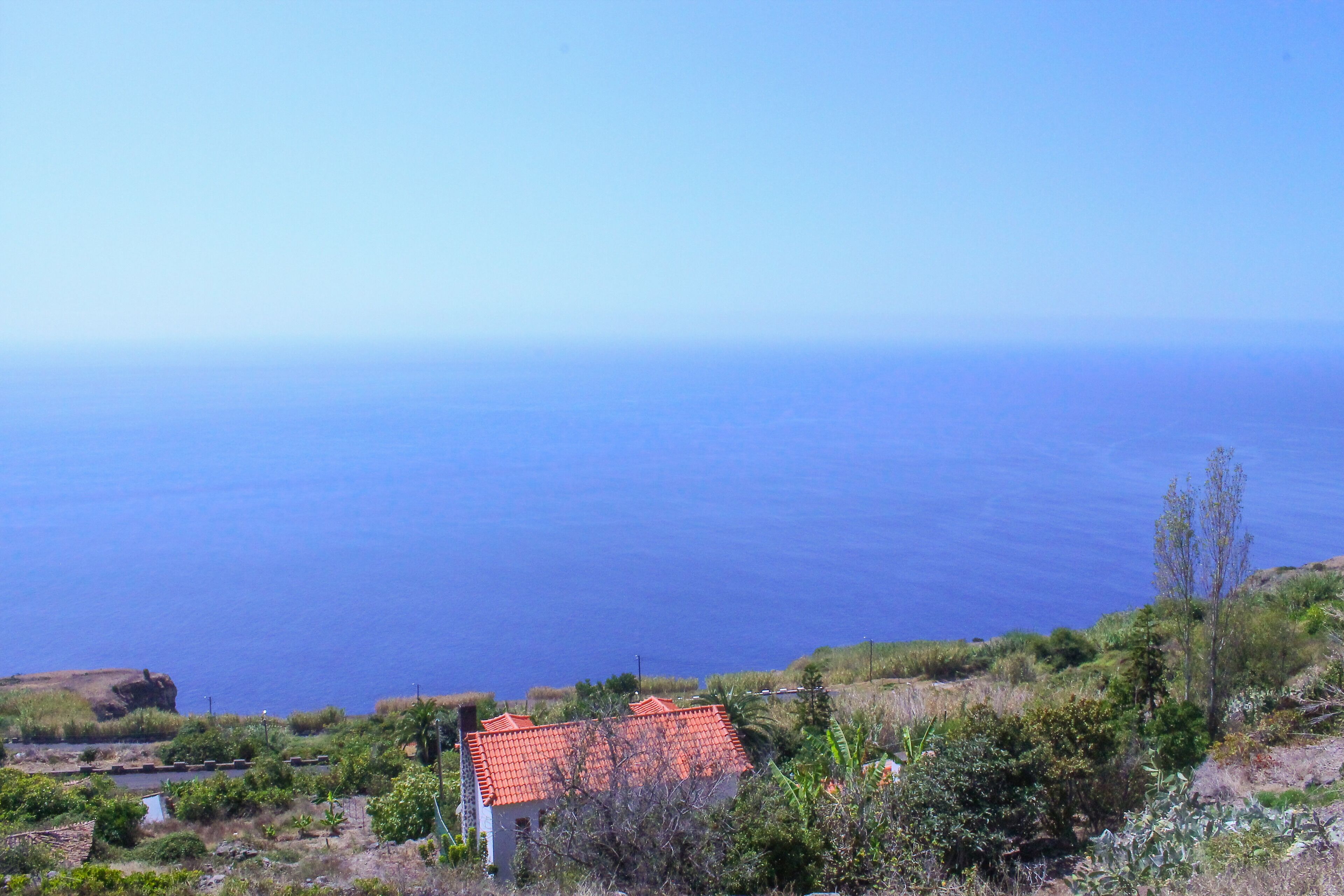 A coastal view with a house featuring a red-tiled roof, surrounded by greenery and overlooking the sea.