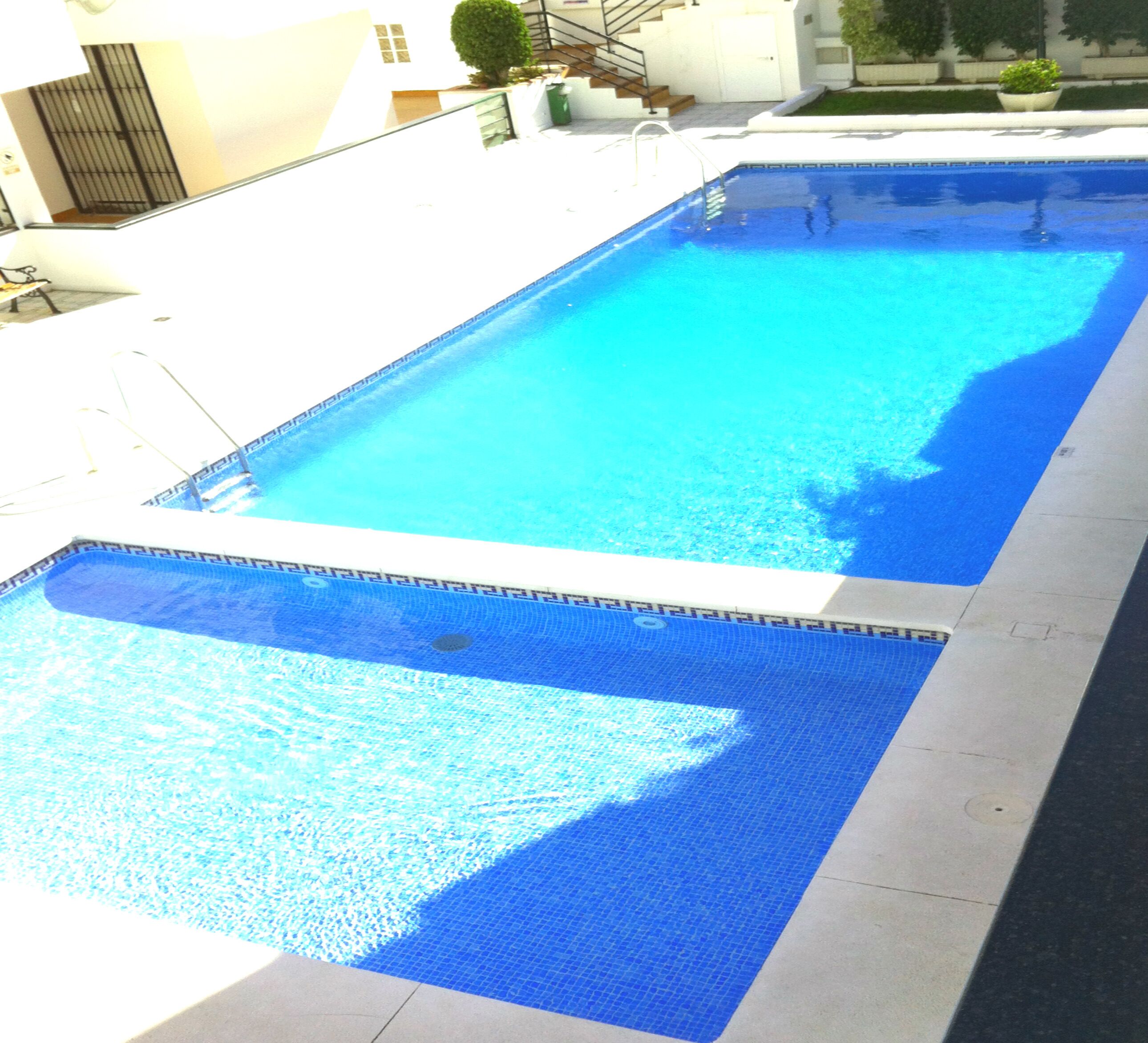 A rectangular swimming pool with a blue mosaic border, surrounded by a white deck and a building in the background.