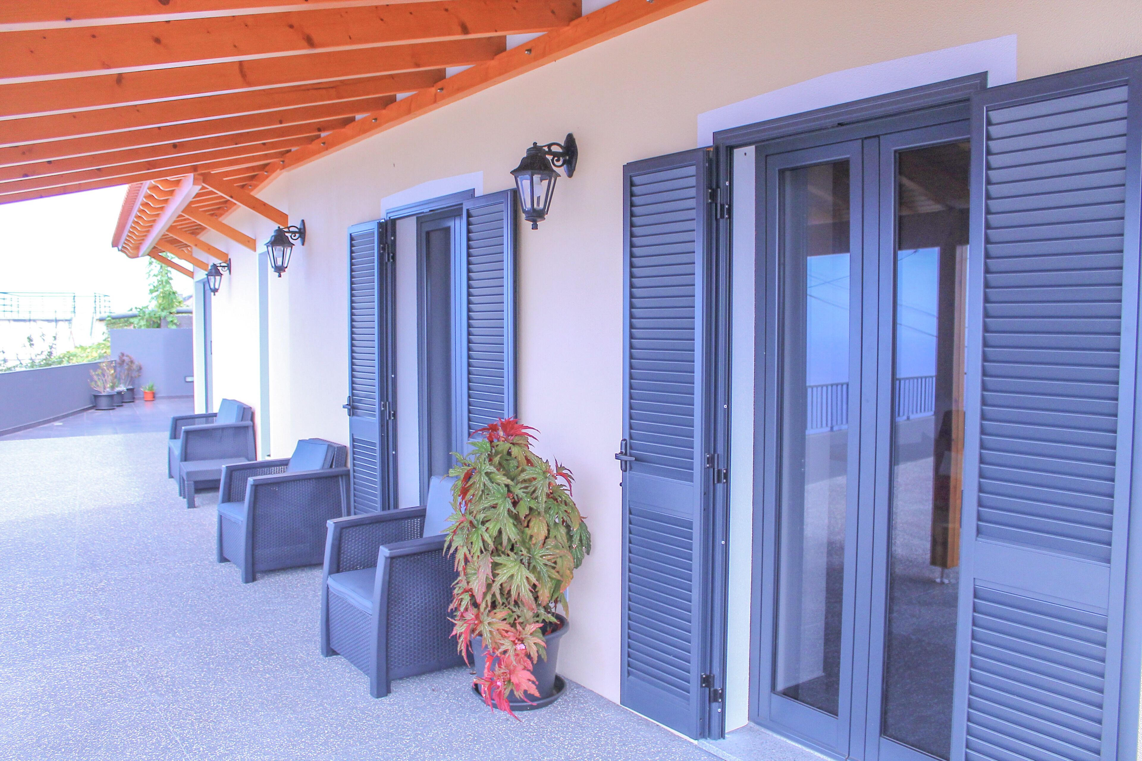 Un espacio de estar techado al aire libre con sillas y una planta en maceta, junto a puertas cerradas con contraventanas.