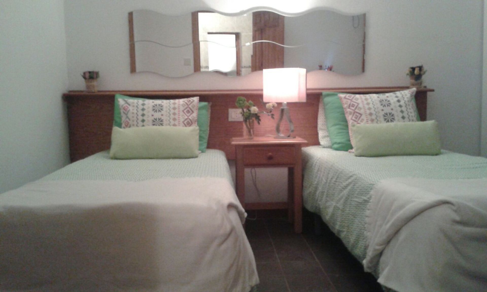 Une chambre d’hôtel avec deux lits simples, une table de chevet avec une lampe, un miroir et une petite table sur laquelle est posé un vase de fleurs.