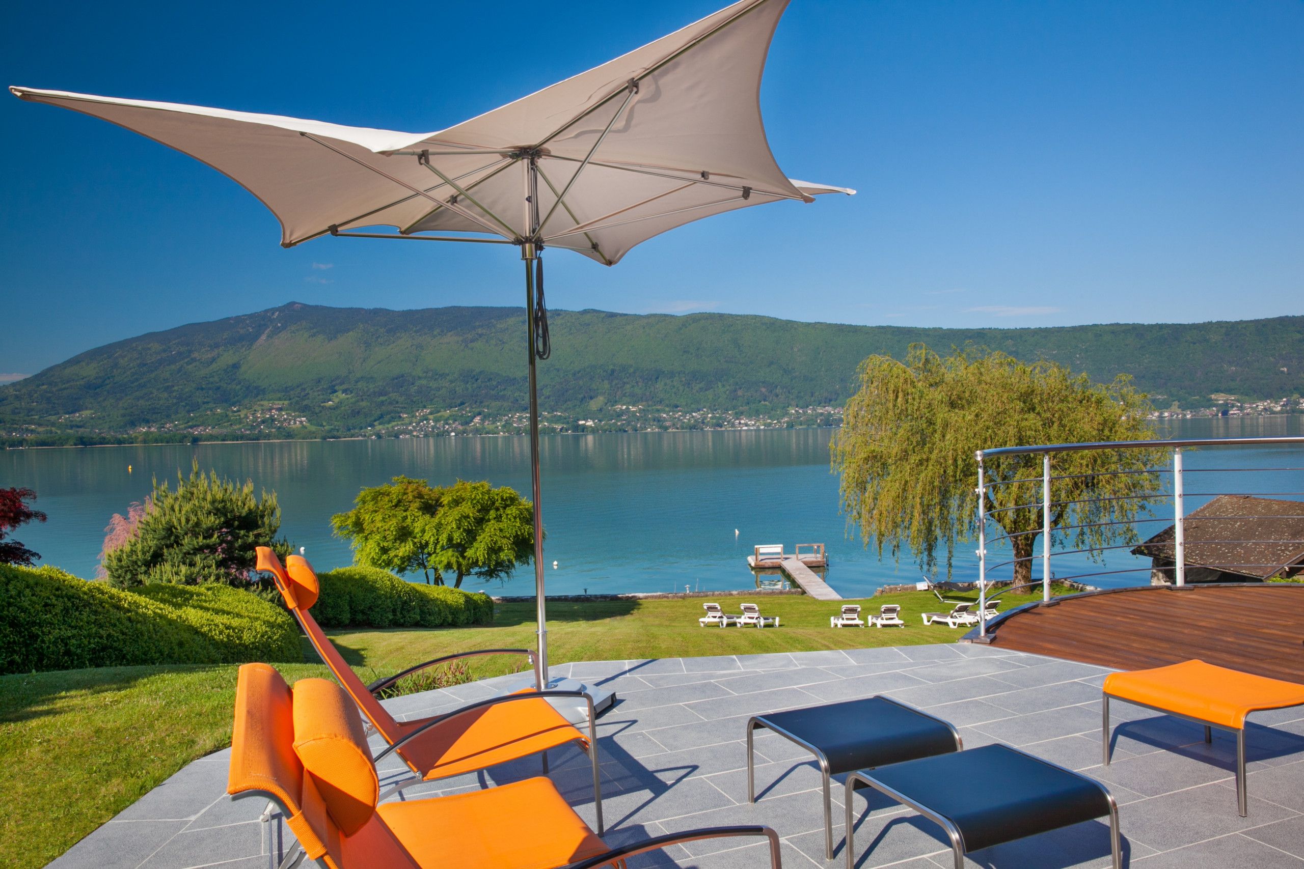 A terrace with outdoor seating, a large umbrella, and a view of a lake with mountains in the background.