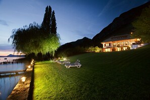 A lakeside property at dusk with a well-lit lawn, lounge chairs, and a large building with multiple windows. - Villa Bagatelle 5 stars Vision Luxe 10 people 5 bedrooms Annecy (Veyrier-du-Lac)