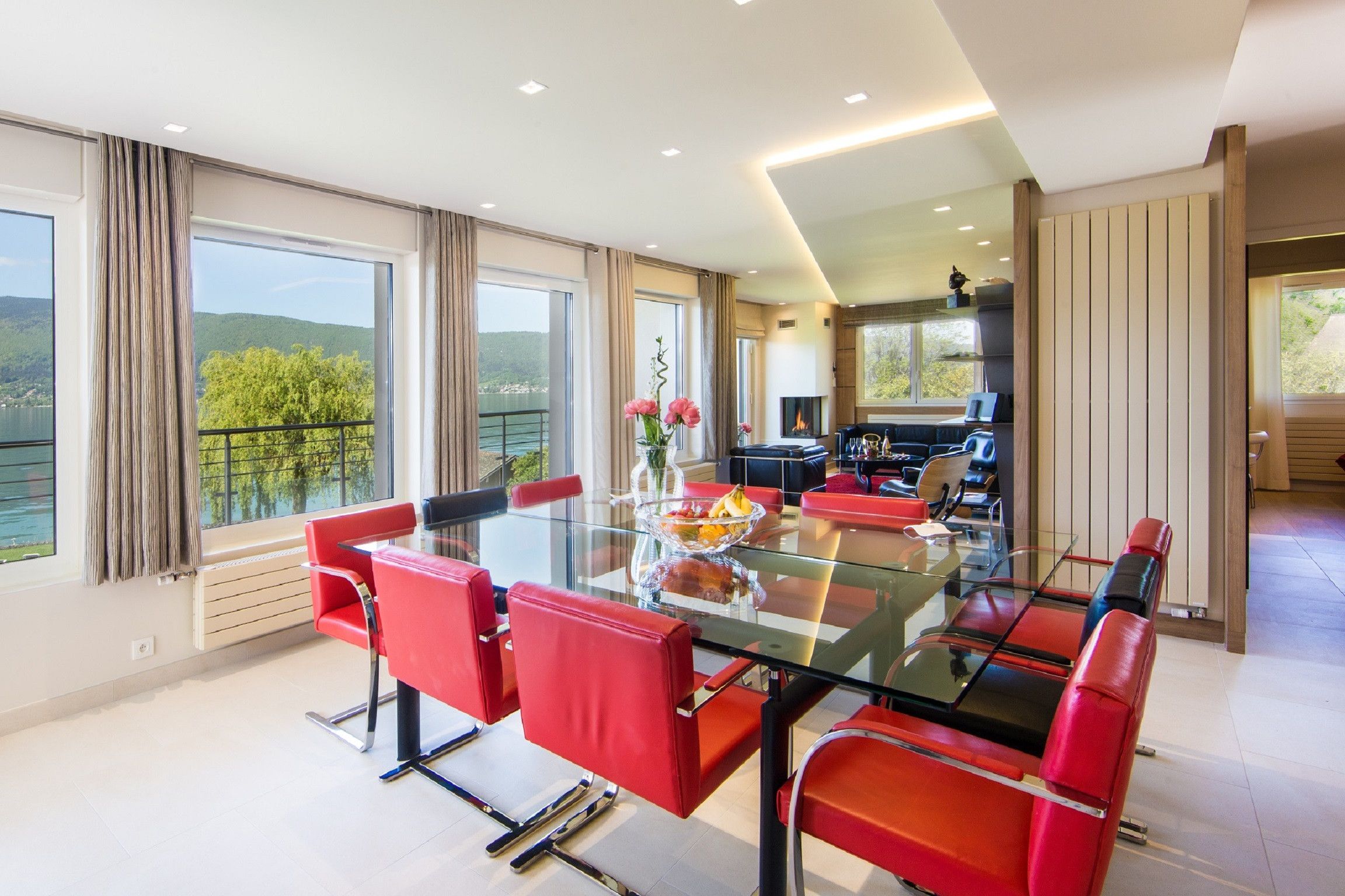 Un espace de repas moderne avec une table en verre et des chaises rouges, de grandes fenêtres avec des rideaux et une vue sur la verdure.