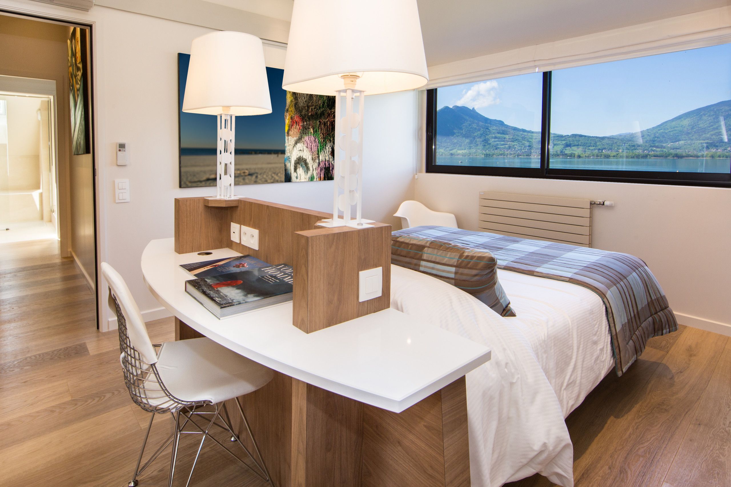 A modern hotel room with a bed, a desk with a chair, a lamp, and a view of a lake and mountains.