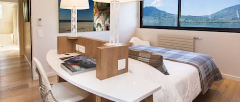 A modern hotel room with a bed, a desk with a chair, a lamp, and a view of a lake and mountains.