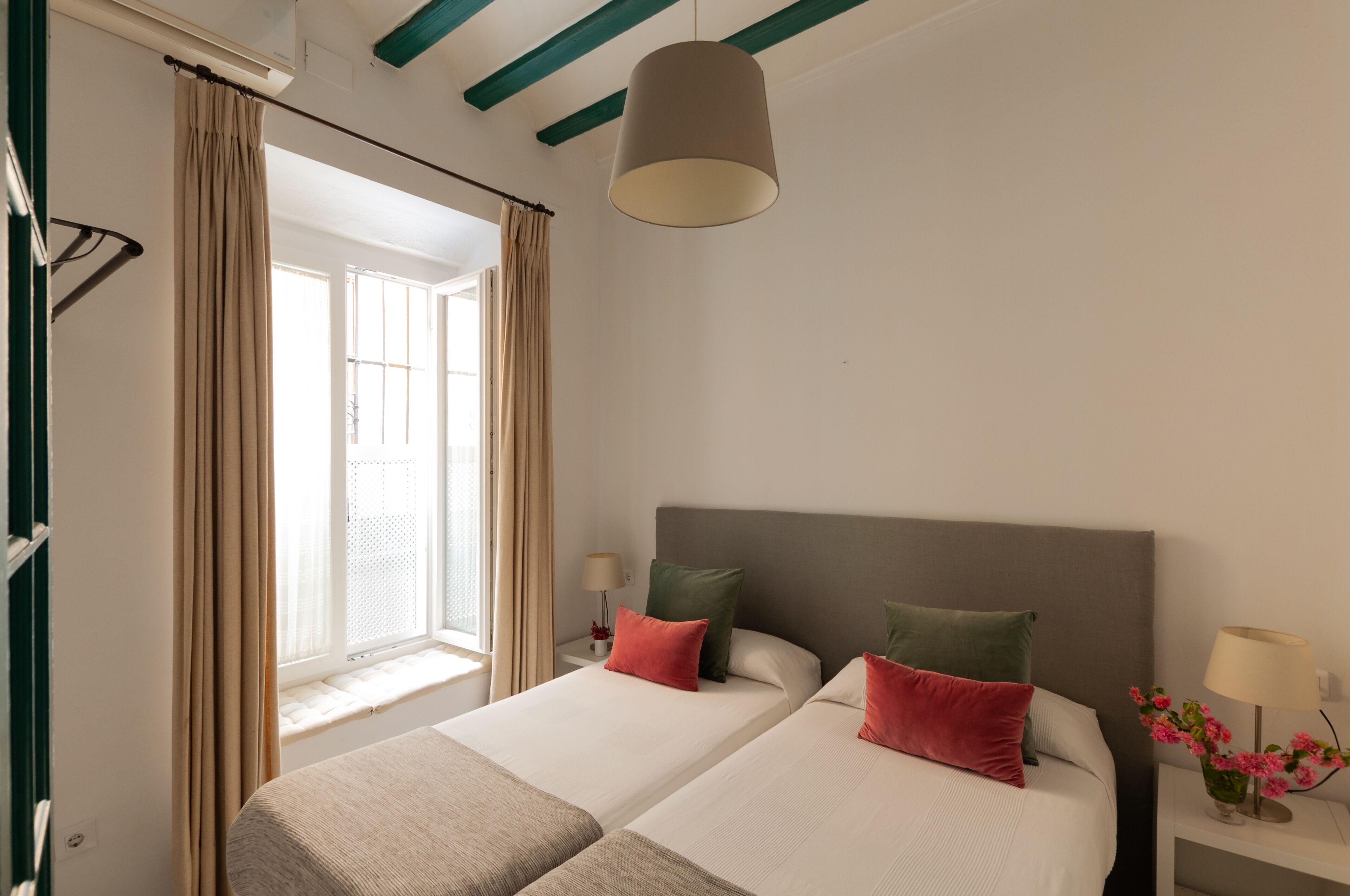 Ein Hotelzimmer mit zwei Betten, einem Fenster mit Vorhängen, einer Hängelampe und einem Nachttisch mit Lampe und Blumen.