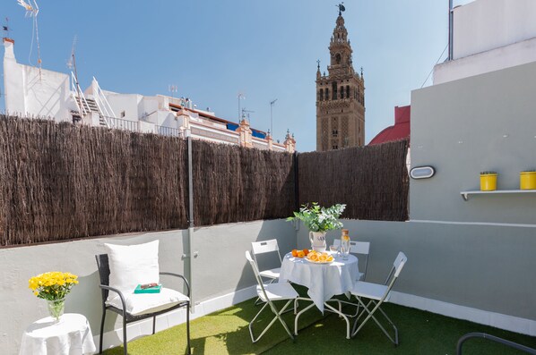 A rooftop terrace with a table set for two, a chair, a small table with a vase of flowers, and a view of a historic tower in the background.