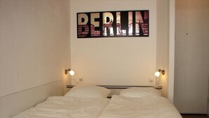 Ein Doppelbett mit weißer Bettwäsche in einem Zimmer, das ein an der Wand befestigtes Schild mit dem Schriftzug „BERLIN“ und zwei Nachttischlampen zeigt.