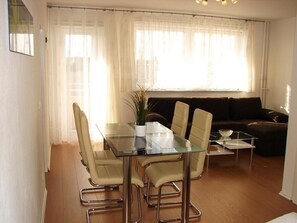 A dining area with a glass table and four chairs, a sofa, and a coffee table in a well-lit room with large windows.