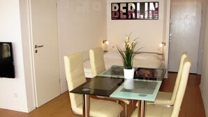 Ein Essbereich mit einem Glastisch, vier beigefarbenen Stühlen, einer Topfpflanze und einem an der Wand befestigten Schild mit der Aufschrift 'BERLIN'.