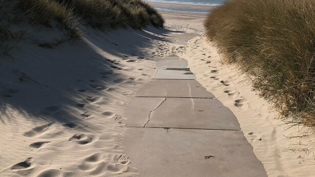 Ein sandiger Küstenpfad führt zum Meer, begleitet von Fußspuren und Gras an beiden Seiten.
