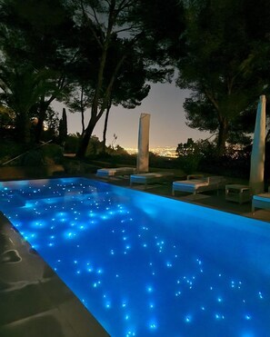 Una piscina exterior con luces azules iluminadas, rodeada de árboles y sillas de descanso.