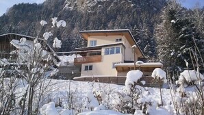 A modern two-story mountain house with a sloped roof, surrounded by snow-covered trees and a rocky mountain backdrop.