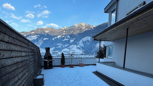 A snow-covered courtyard with a wooden fence, potted plants, and a view of snow-capped mountains in the background.