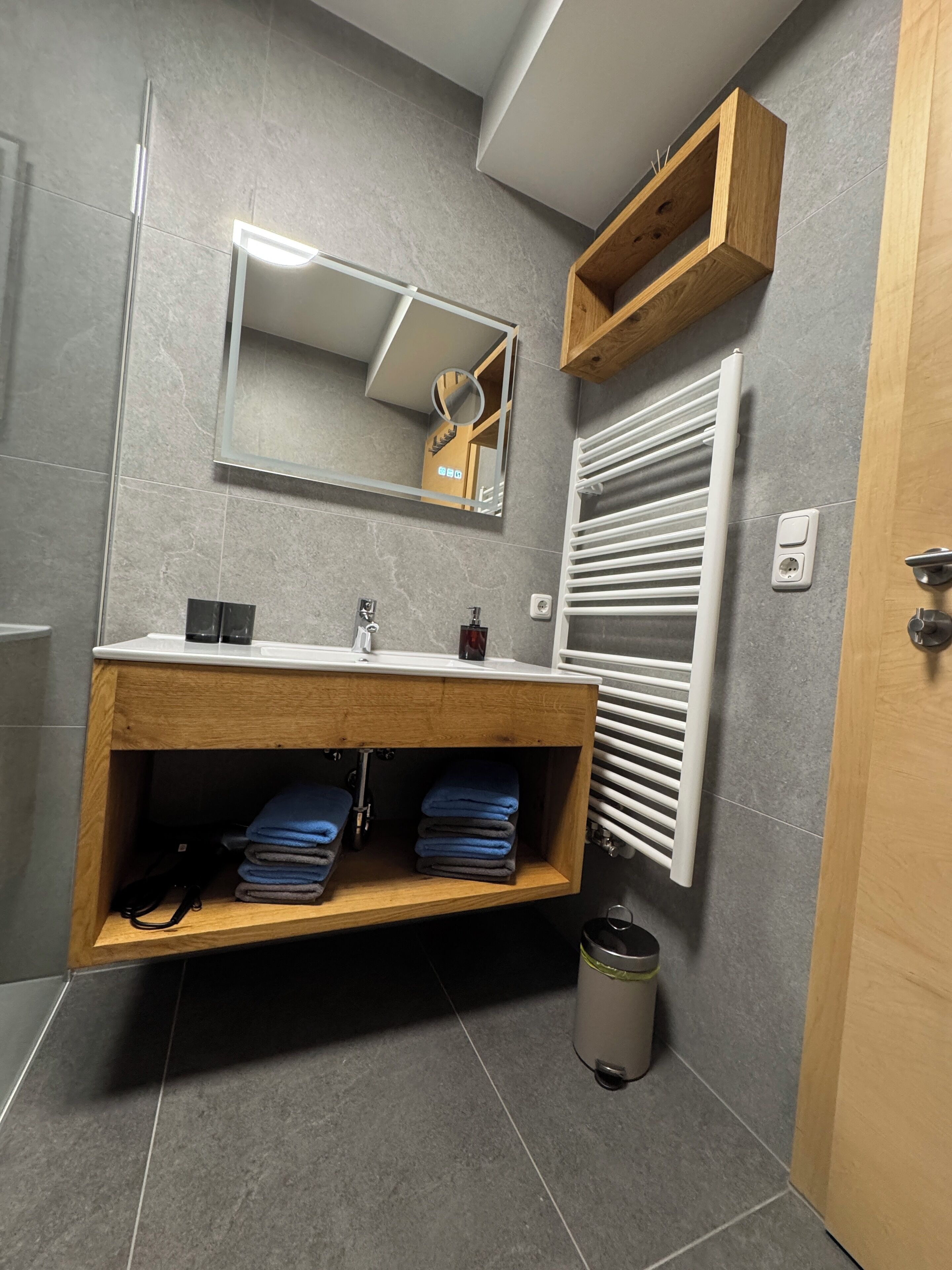 A modern bathroom with grey tiles, a wooden vanity with a mirror, a towel rack, and a radiator.
