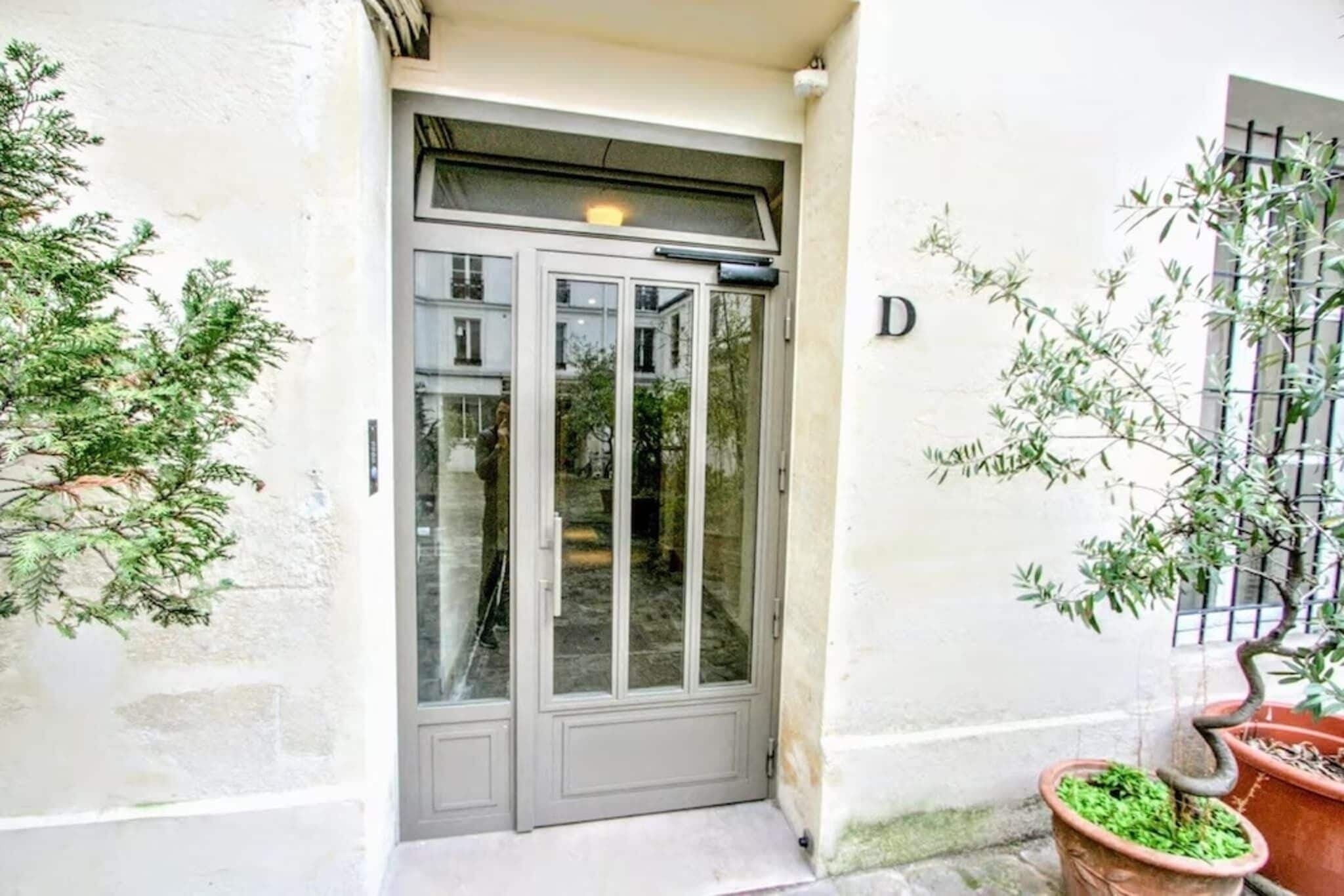 Una puerta doble gris con un panel de vidrio, una pequeña ventana con celosía metálica y una planta en maceta a la derecha.
