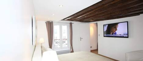 A bedroom with a bed, a TV mounted on the wall, a window with curtains, and a wooden ceiling.