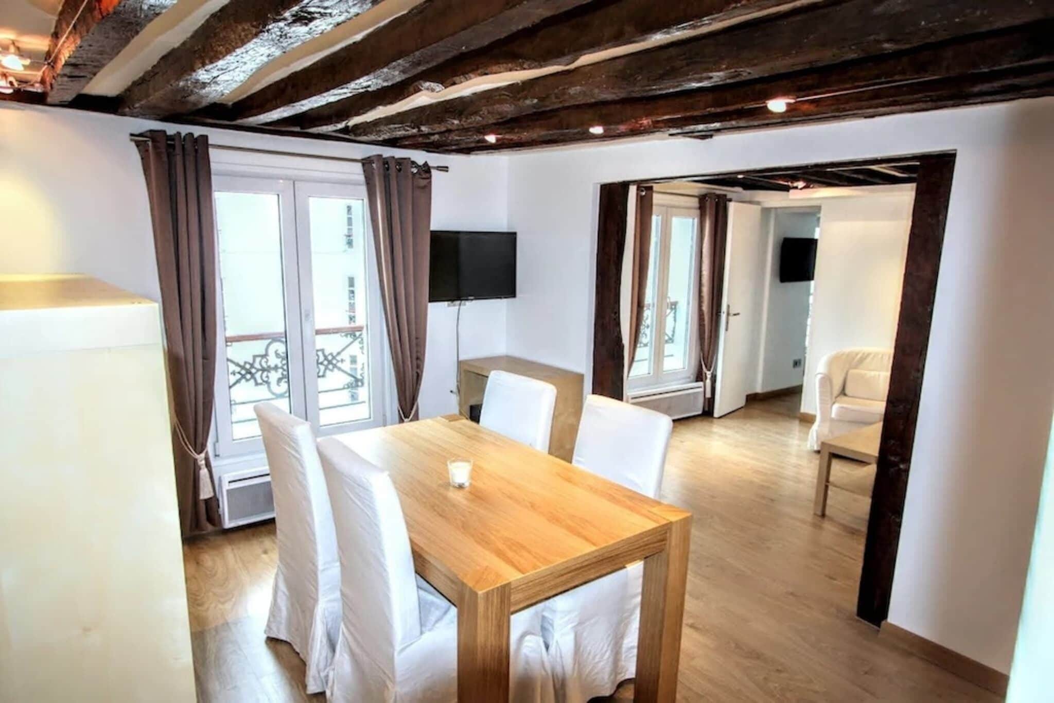Una habitación con una mesa de comedor de madera, sillas blancas, un televisor montado en la pared y un balcón con cortinas.