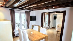 A room with a wooden dining table, white chairs, a TV mounted on the wall, and a balcony with curtains. - Superb 4-room apartment (Paris)
