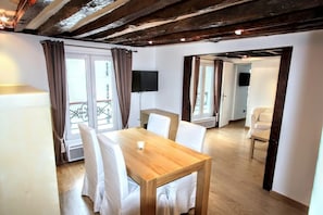 Una habitación con una mesa de comedor de madera, sillas blancas, un televisor montado en la pared y un balcón con cortinas.