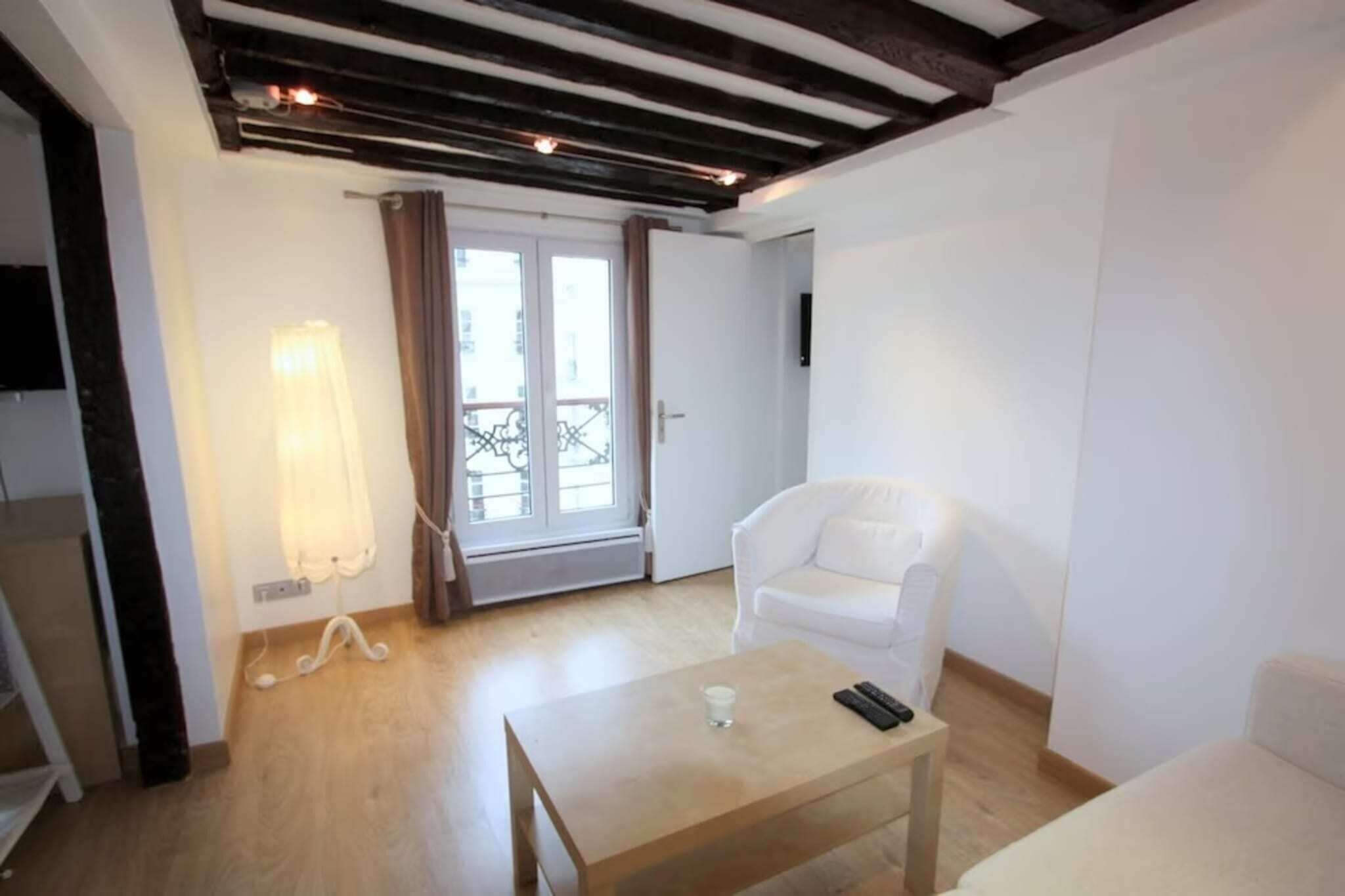 Quarto com piso de madeira, um sofá branco, uma mesa pequena com um copo e um controle remoto, e uma porta que dá para uma varanda.