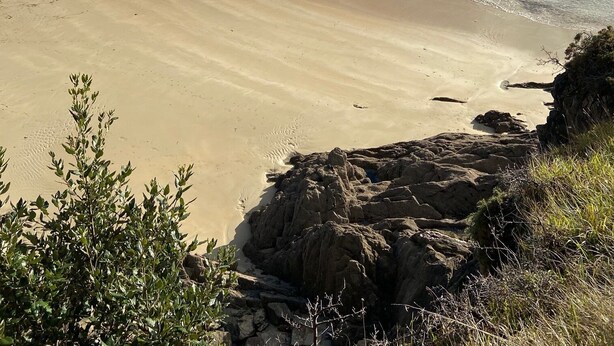 Une plage côtière avec un rivage sablonneux, des affleurements rocheux et quelques maisons en arrière-plan.