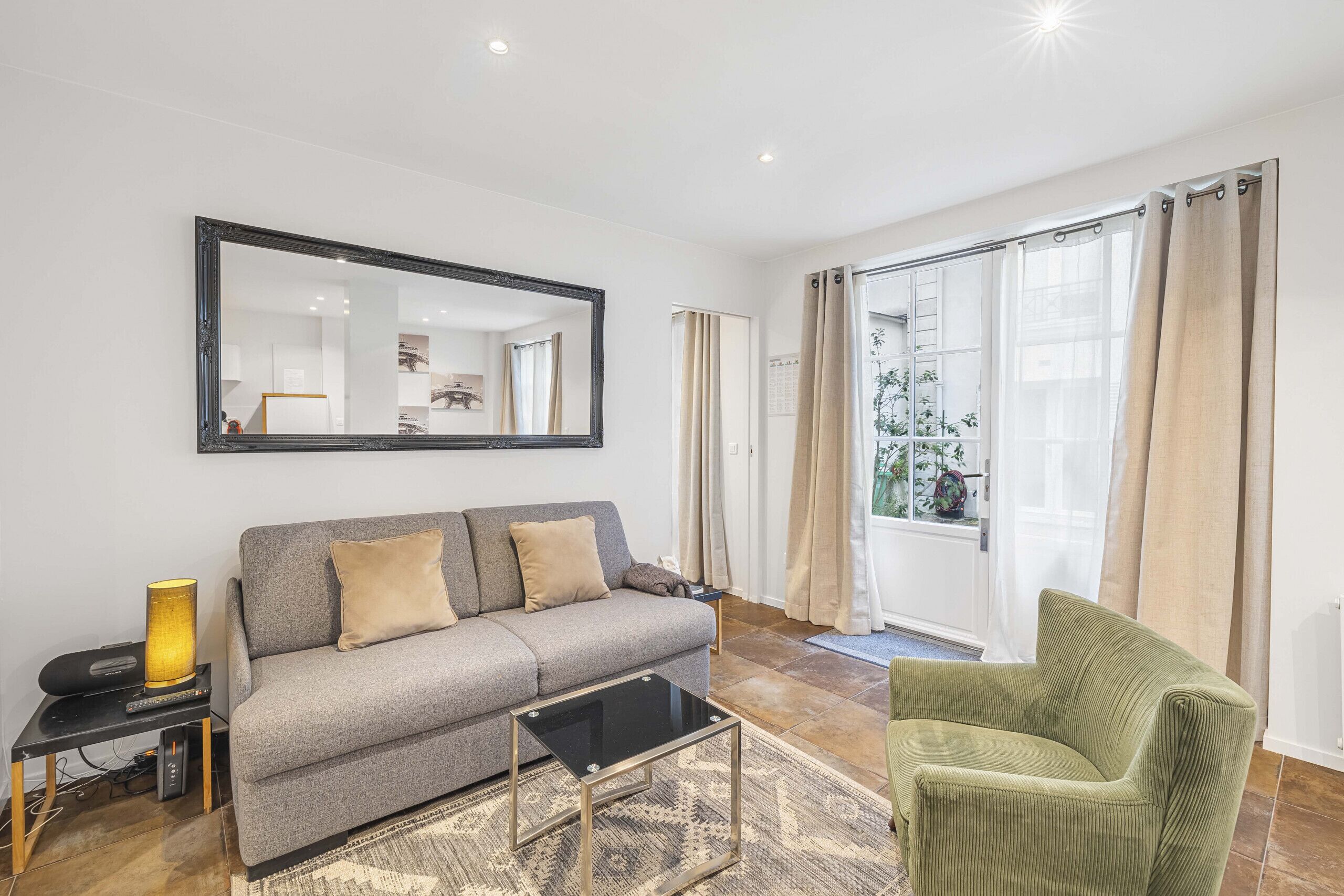 A living room with a grey sofa, a green armchair, a black coffee table, a large mirror, and a window with curtains.