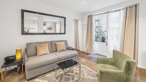 Un salon avec un canapé gris, un fauteuil vert, une table basse noire, un grand miroir et une fenêtre avec des rideaux.