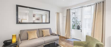A living room with a grey sofa, a green armchair, a black coffee table, a large mirror, and a window with curtains.