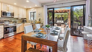 Una cocina moderna con electrodomésticos de acero inoxidable, una zona de comedor con una mesa y sillas de madera, y un amplio ventanal corredero que da a un patio exterior.