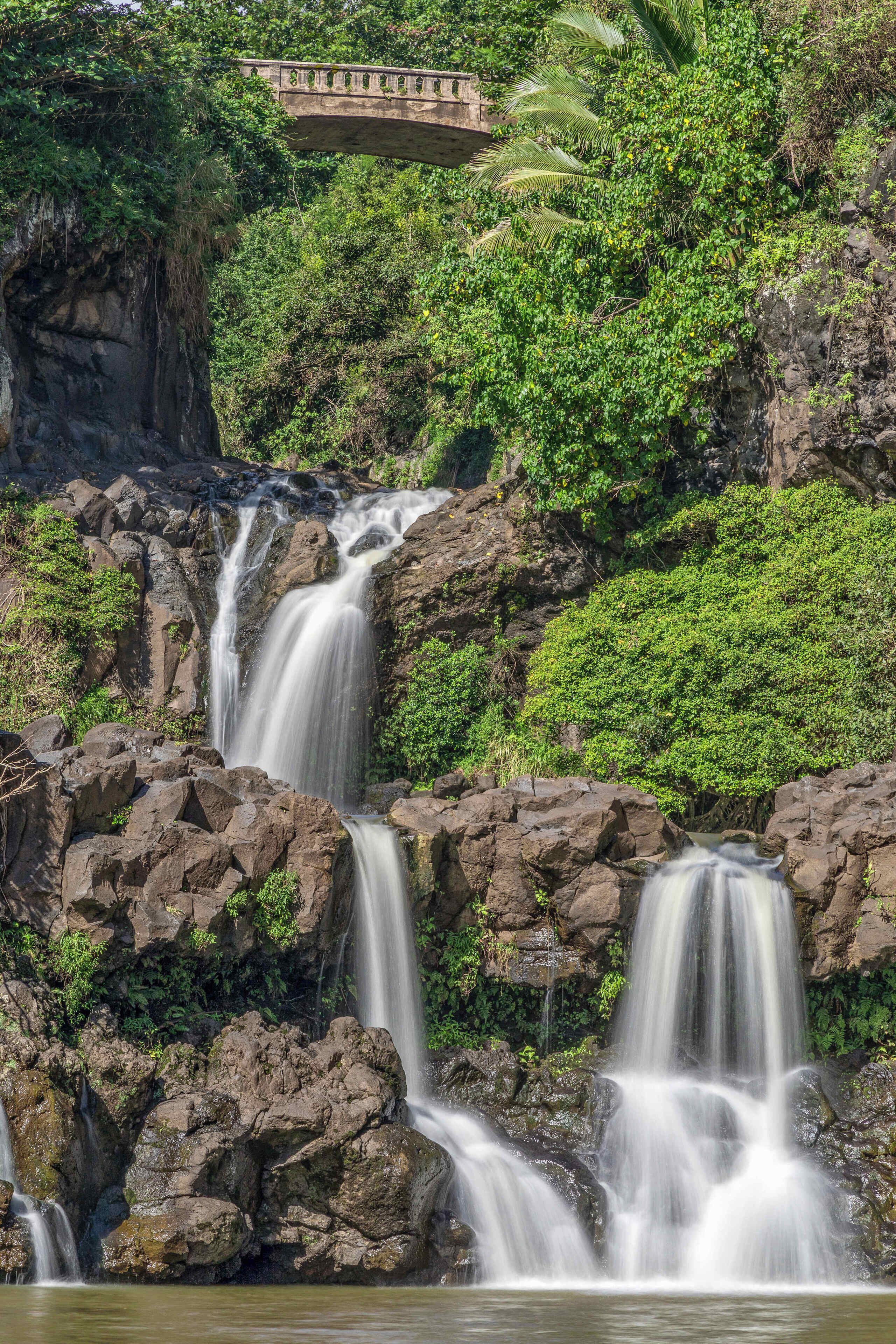 Una cascata che si getta su rocce, con vegetazione rigogliosa e un ponte sullo sfondo.