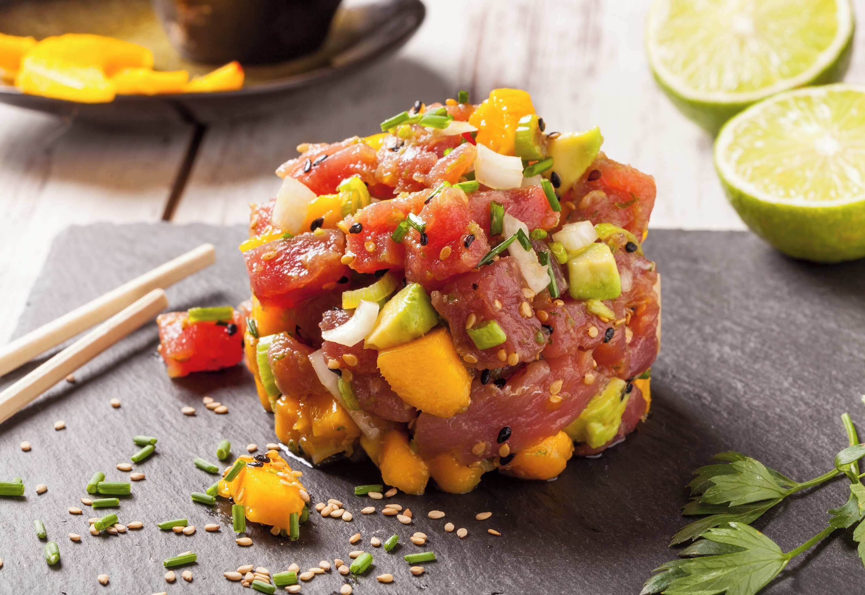Un plat de tartare de thon cru, coupé en dés, accompagné de mangue et d’oignons, garni de graines de sésame et d’oignons verts, servi sur une ardoise.