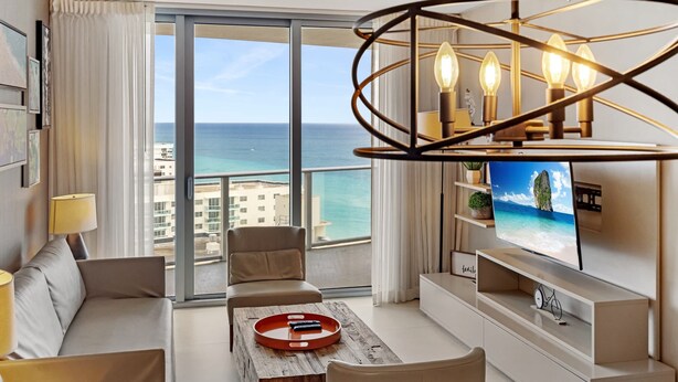 A modern living room with a sofa, coffee table, and TV. A large window offers a view of the ocean and buildings.