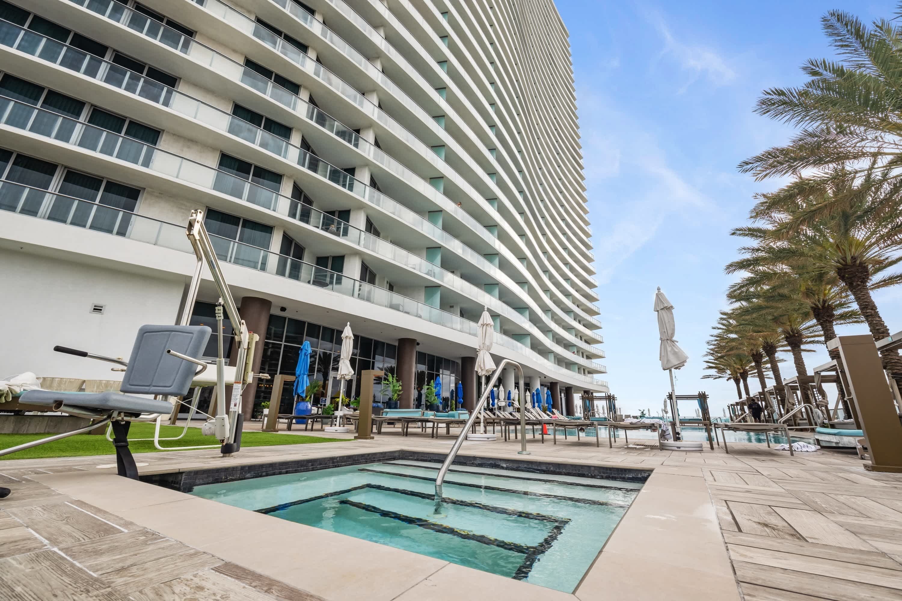 A modern hotel with a pool area, including lounge chairs and umbrellas, and a tall building in the background.